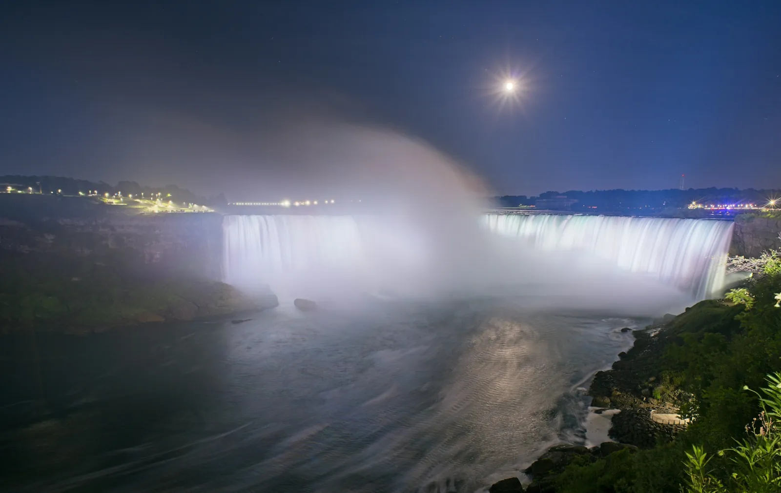 Horseshoe Falls glowing in floodlight under a bright moon, with a plume of mist rising from the gorge into a deep blue sky